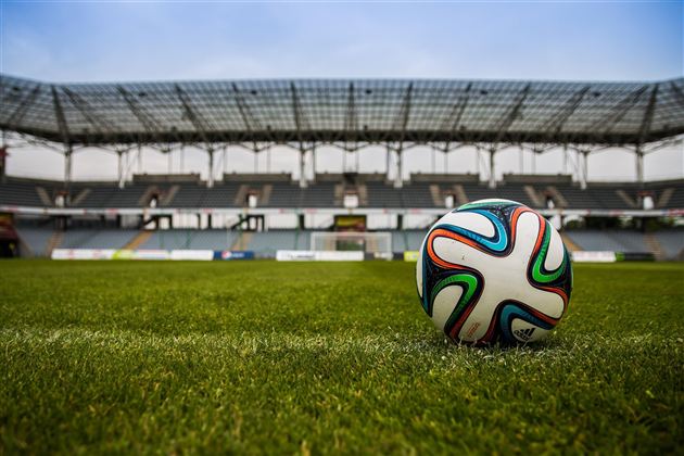 Fotbalový míč na zelené trávě před prázdným stadionem. Obloha je modrá a tribuny jsou viditelné.
