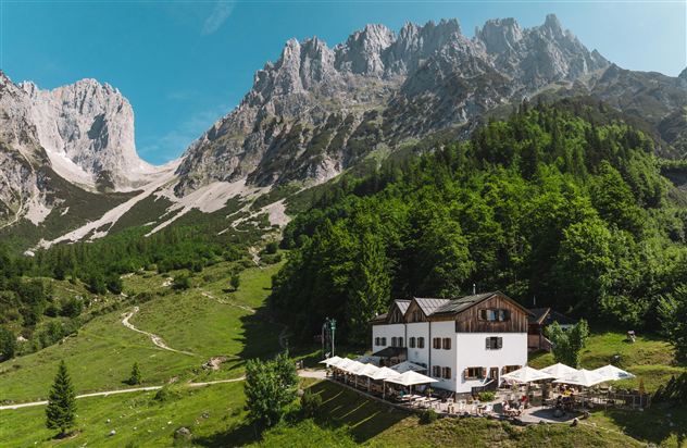 Eine malerische Berghütte umgeben von hohen Bergen und grünen Wäldern. Auf der Terrasse stehen Tische mit Sonnenschirmen.
