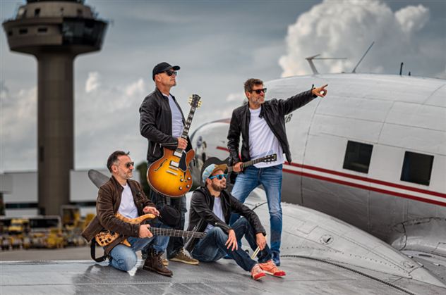 A band poses on top of an airplane, surrounded by clouds and an airport building. The musicians are holding guitars and wearing casual clothing.