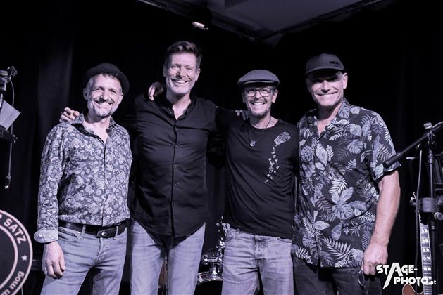 Eine Rockband posiert auf dem Dach eines Flugzeugs. Im Hintergrund sieht man einen Flugturm und einen bewölkten Himmel.