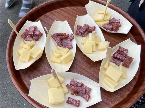 Ein Holztablett mit sechs kleinen Schalen, gefüllt mit Käse und Wurststücken. Die Häppchen sind an Holzstäbchen befestigt.