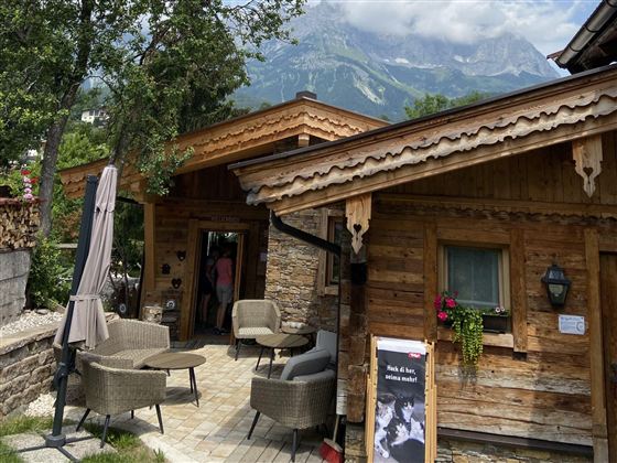 Ein gemütliches Holzhaus in den Bergen mit Terrassenmöbeln und Bäumen. Im Hintergrund sind große Berge und ein bewölkter Himmel zu sehen.