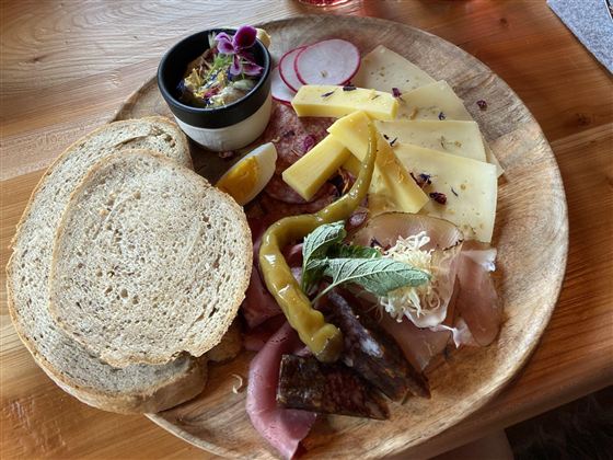 Eine rustikale Platte mit verschiedenen Käsesorten, Wurst, Brot und frischem Gemüse. Dazu gibt es einen kleinen Dip in einer Schüssel.