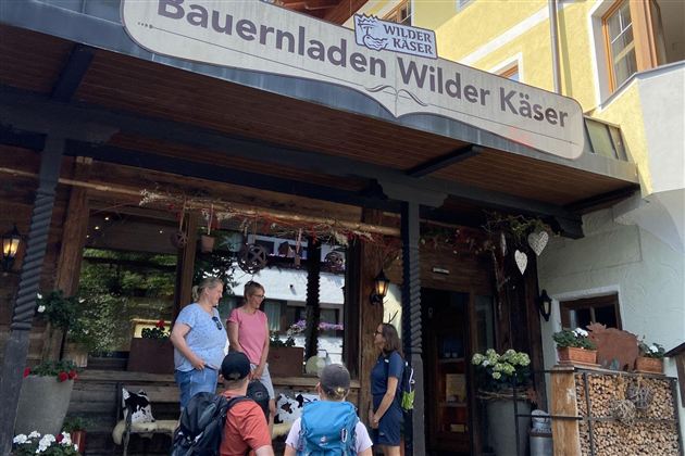 Ein Bauernladen mit dem Namen "Wilder Käser" ist zu sehen. Einige Personen stehen vor dem Eingang und unterhalten sich.