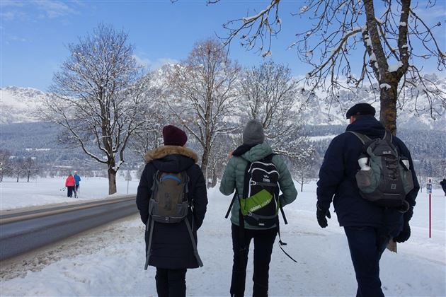 Drie personen wandelen op een sneeuwbedekte weg. Op de achtergrond zijn besneeuwde bergen en bomen te zien.