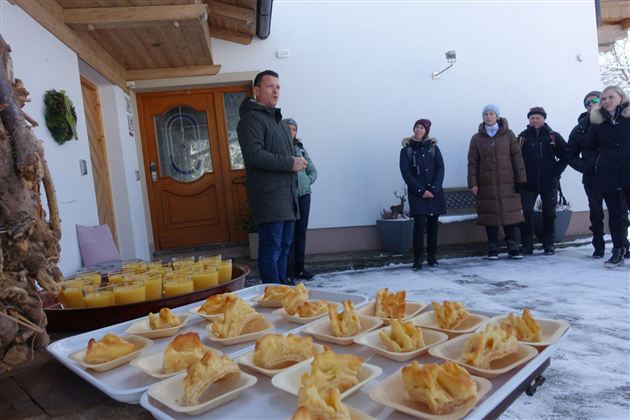 Eine Gruppe von Menschen steht vor einem Gebäude und hört einem Sprecher zu. Auf einem Tisch vor ihnen sind mehrere Teller mit Essen angerichtet.