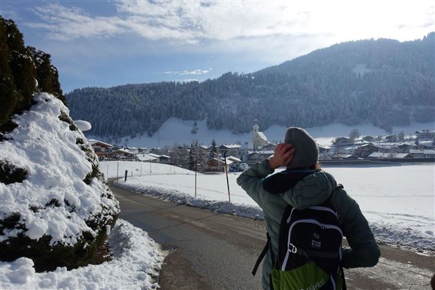 Een persoon staat aan een besneeuwde straat en kijkt naar de bergen. Op de achtergrond zijn houten gebouwen en een kerktoren te zien.