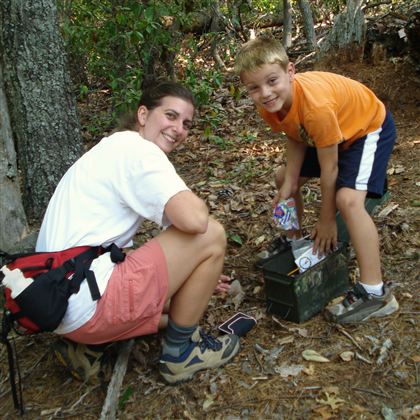En kvinde og en dreng leder efter en skat i en skov. De smiler og kigger i en kasse, der er gemt i jorden.