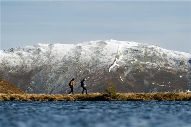Two hikers by the shore of a lake in front of snow-covered mountains. The sky is clear and the scenery is picturesque.