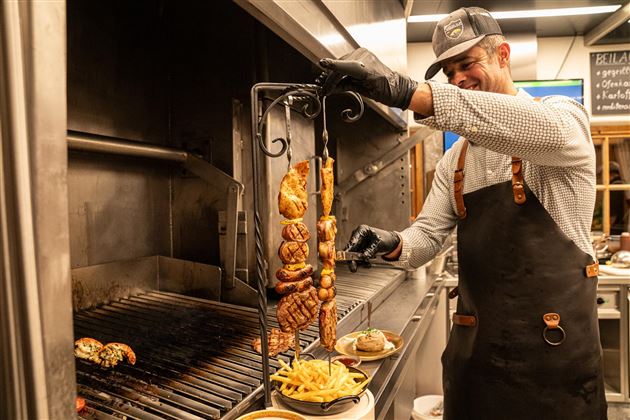 Ein Koch steht am Grill und bereitet Fleisch auf einem Spieß zu. Im Vordergrund sind Pommes frites und ein Burger zu sehen.