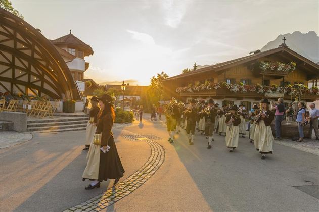 Eine Gruppe von Menschen in traditioneller Kleidung marschiert durch eine malerische Straße. Im Hintergrund sind schöne Holzhäuser und eine sonnige Atmosphäre sichtbar.