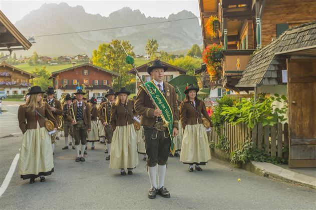 Eine festliche Parade mit einer Gruppe von Menschen in traditioneller Tracht. Im Hintergrund sind malerische Berge und Holzhäuser zu sehen.
