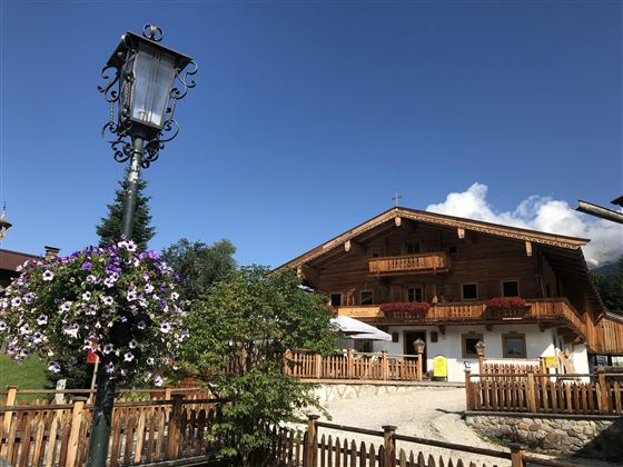 Ein traditionelles Holzhaus mit Balkon und schönen Blumen vor der Tür. Im Hintergrund ist ein blauer Himmel mit einigen Wolken zu sehen.