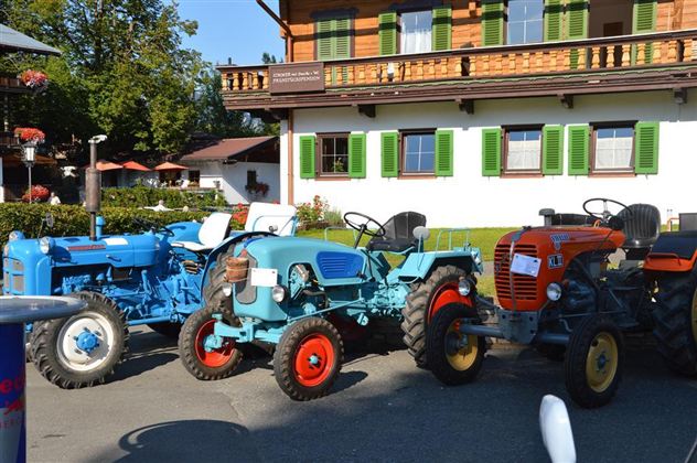 Drei bunte Traktoren stehen vor einem traditionellen bayerischen Gebäude. Der Himmel ist klar und die Umgebung ist grün und einladend.