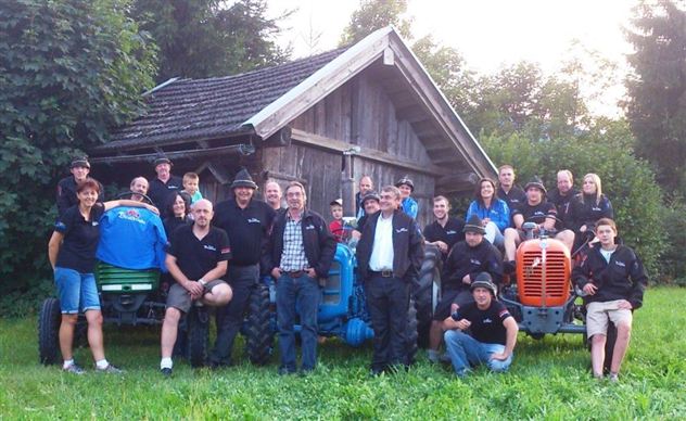Eine Gruppe von Menschen steht vor einer alten Scheune mit Traktoren. Alle tragen schwarze T-Shirts und lächeln in die Kamera.