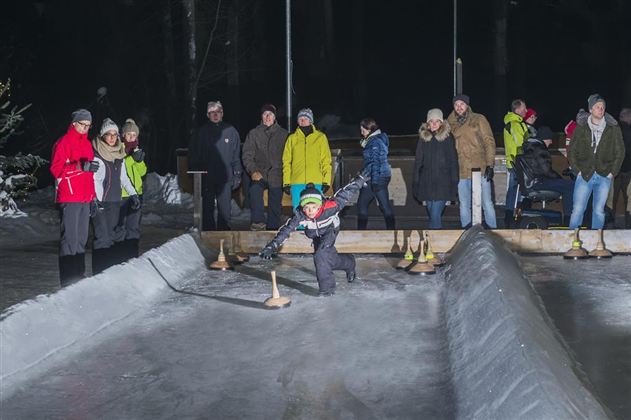 Eine Gruppe von Menschen steht am Rand einer Eisbahn, während ein Kind mit einem Stock auf dem Eis spielt. Es ist eine kalte Winternacht und die Zuschauer sind warm gekleidet.