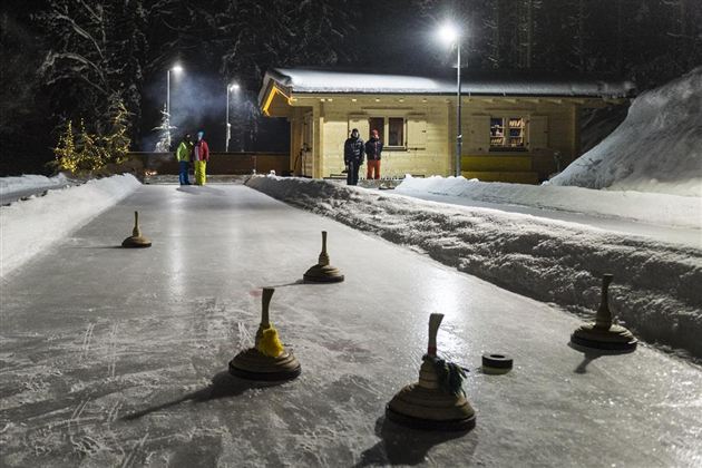 Eine Eisbahn, beleuchtet von Laternen, mit fünf Holzstöcken auf dem Eis. Im Hintergrund stehen mehrere Personen und eine kleine Hütte.