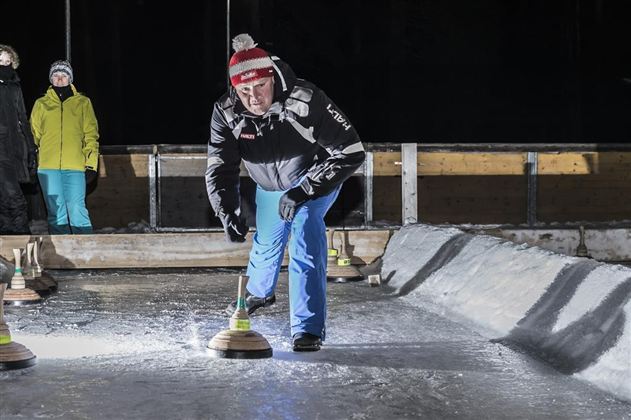 Ein Mann spielt Eisstockschießen auf einer beleuchteten Bahn. Im Hintergrund sind Zuschauer in Winterkleidung zu sehen.