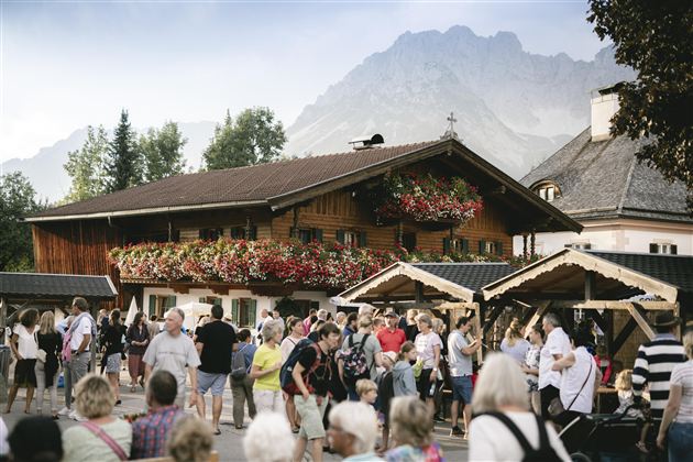 Ein belebter Marktplatz mit vielen Menschen vor einem traditionellen Holzhaus. Im Hintergrund sind Berge und Bäume zu sehen, die eine idyllische Atmosphäre schaffen.