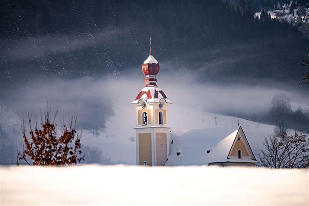 Ein Kirchturm mit einer roten Kuppel ragt aus einer schneebedeckten Landschaft hervor. Im Hintergrund sind verschneite Hügel und Nebel zu sehen.