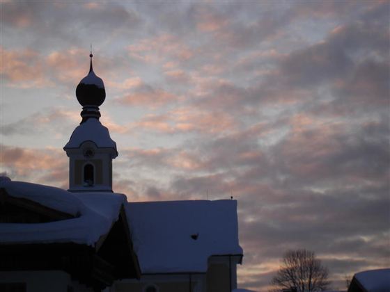 Een sneeuwlandschap met een kerktoren, die tegen de avondlucht is afgebeeld. De lucht is in zachte kleuren gekleurd, wat een rustige sfeer creëert.