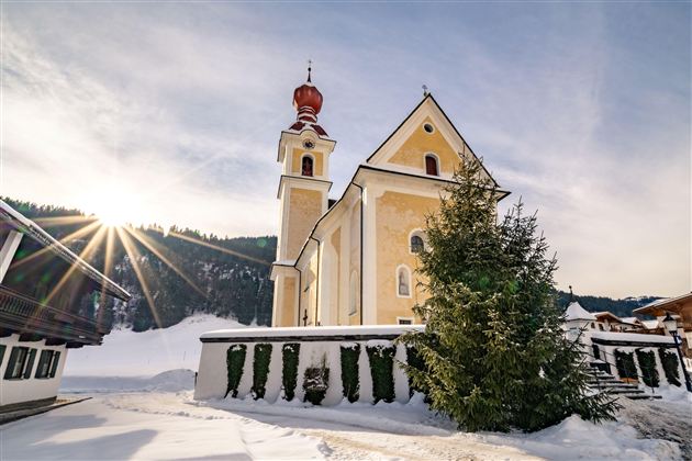 Eine schöne Kirche in einer schneebedeckten Landschaft. Die Sonne scheint hinter den Bergen und beleuchtet die Umgebung.