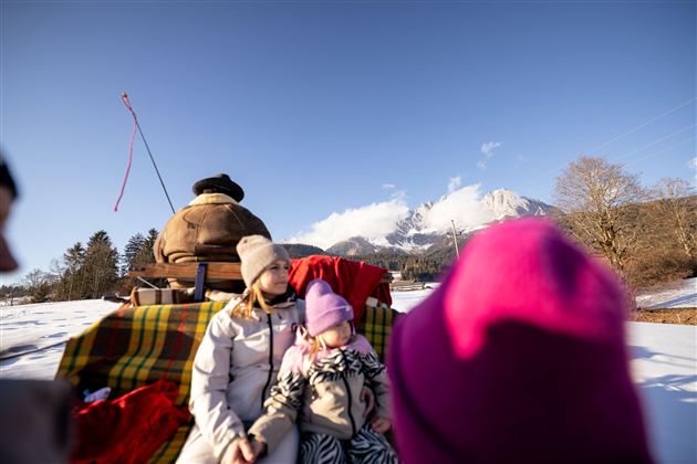 Eine Familie sitzt auf einem Schlitten im Schnee, umgeben von einer winterlichen Landschaft. Im Hintergrund sind Berge unter einem klaren blauen Himmel zu sehen.
