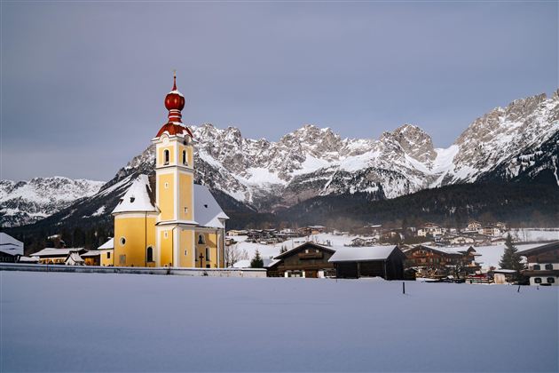 Eine bunte Kirche steht in einer verschneiten Landschaft. Im Hintergrund erheben sich majestätische Berge.