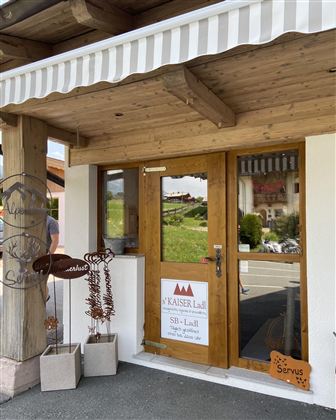 Der Eingang zu einem charmanten Geschäft mit Holzverkleidung und einem weißen Sonnenschirm. Auf der Tür hängt ein Schild mit dem Namen "Kaiser Ladl".