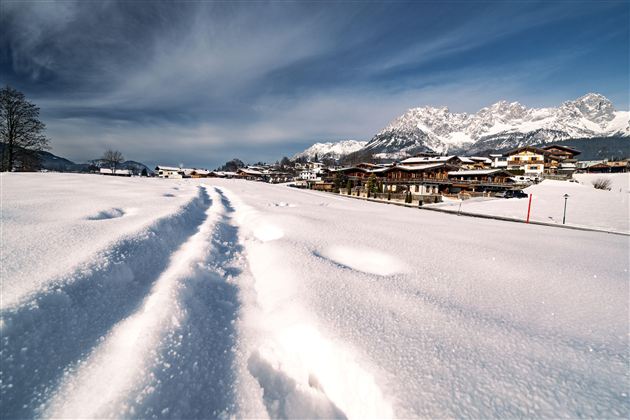 Eine schneebedeckte Landschaft mit klaren Fußspuren im Schnee. Im Hintergrund sind Berge und ein schöner Ort zu sehen.