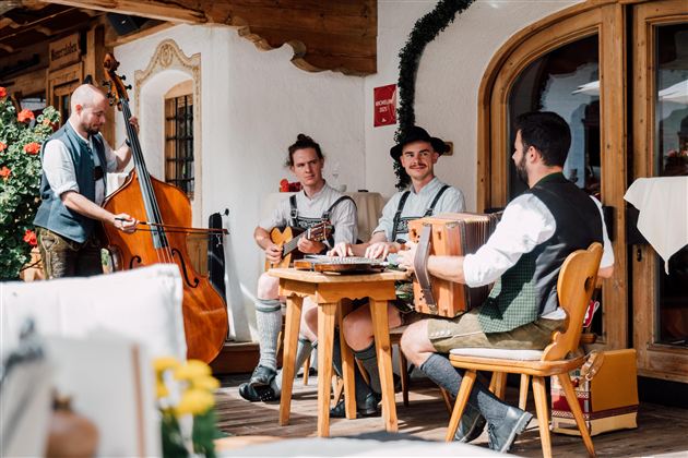 Eine Gruppe von Musikern spielt traditionelle Instrumente in einem gemütlichen Außenbereich. Die Musiker tragen bayerische Trachten und umgeben sich mit schöner Dekoration.