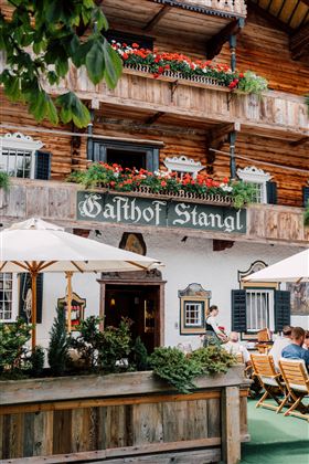 Ein traditionelles Gasthaus mit holzverkleideter Fassade und blühenden Pflanzen. Tische im Freien unter Sonnenschirmen bieten einen einladenden Platz zum Verweilen.