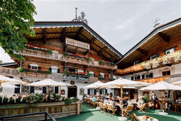 Ein traditionelles alpenländisches Gasthaus mit Holzfassade und blühenden Balkonen. Gäste sitzen unter Sonnenschirmen im Freien und genießen die Atmosphäre.