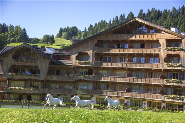 Ein traditionelles Holzgebäude im malerischen Landschaftsraum, umgeben von grünen Wiesen und Wäldern. Vor dem Gebäude laufen stilisierte Pferde im Gras.