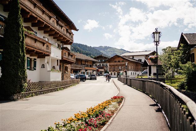 A picturesque street in an alpine town with traditional wooden houses and colorful flower beds. In the background, mountains and a blue sky are visible.