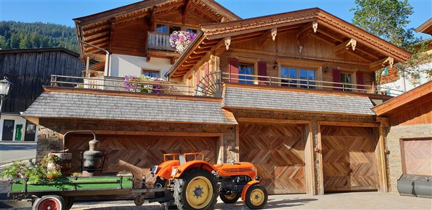 Ein traditionelles Holzhaus mit einem Balkon und Blumen. Vor dem Haus steht ein oranger Traktor und ein Wagen mit Blumen.