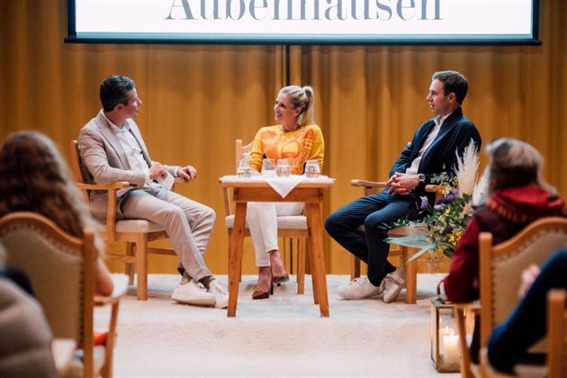Eine Panel-Diskussion mit drei Personen auf einer Bühne. Im Hintergrund ist ein großes Banner mit dem Wort "Außenmalen" zu sehen.