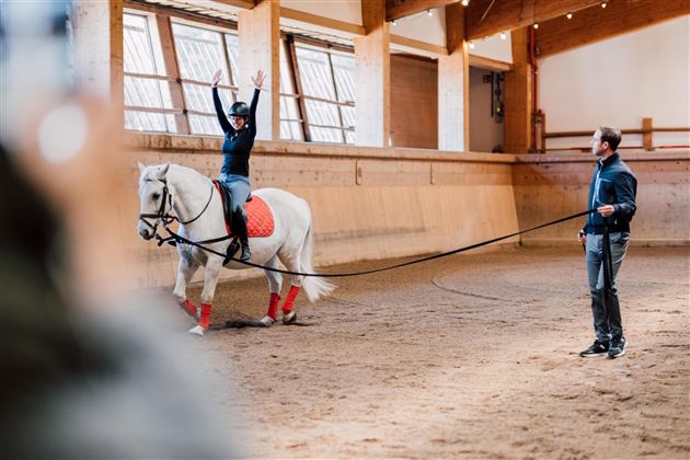 Eine Reiterin zeigt eine Akrobatikübung auf einem weißen Pferd in einer Reitarena. Der Trainer steht daneben und beobachtet die Übung.