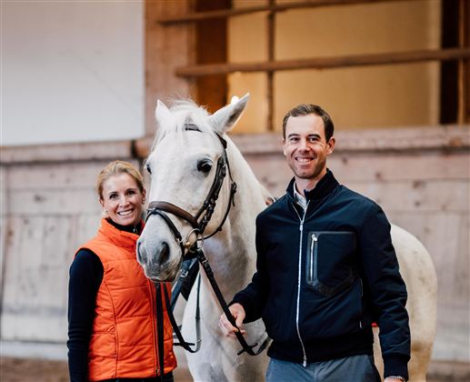 Eine Frau und ein Mann stehen mit einem weißen Pferd in einem Reitstall. Der Mann trägt eine dunkle Jacke und die Frau eine orange Weste.