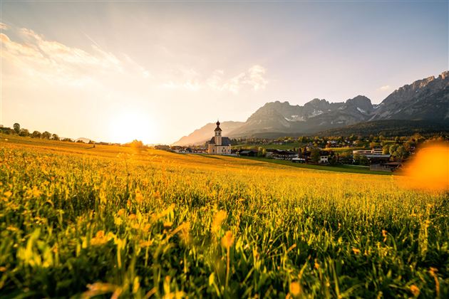 A picturesque landscape with a meadow full of flowers and majestic mountains in the background. The sun sets behind the church, creating a warm atmosphere.