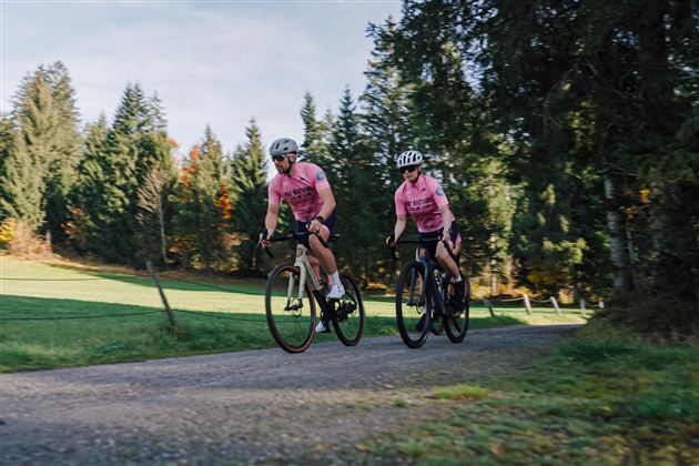 Zwei Radfahrer in rosa Trikots fahren auf einem Schotterweg. Im Hintergrund sind grüne Wiesen und Bäume zu sehen.