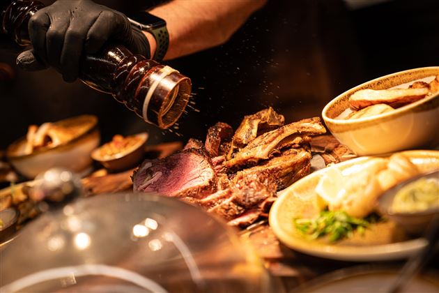 Ein köstliches Stück Fleisch wird frisch gewürzt. Dazu liegen Beilagen wie Kartoffeln und Gemüse bereit.