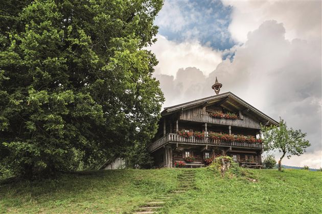 Ein traditionelles Holzhaus steht auf einem sanften Hügel, umgeben von Bäumen. Der Himmel ist bewölkt, was eine ruhige Atmosphäre schafft.