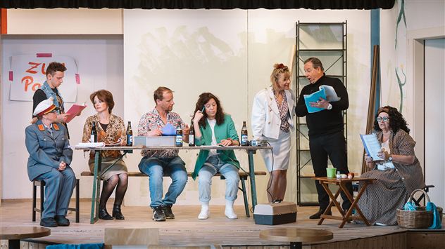 A theater performance with several actors on stage. The performers are in vibrant costumes and interact at a table.