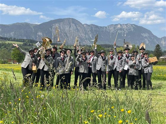 A group of musicians in gray suits stands on a meadow with mountains in the background. They hold instruments and pose happily for the photo.