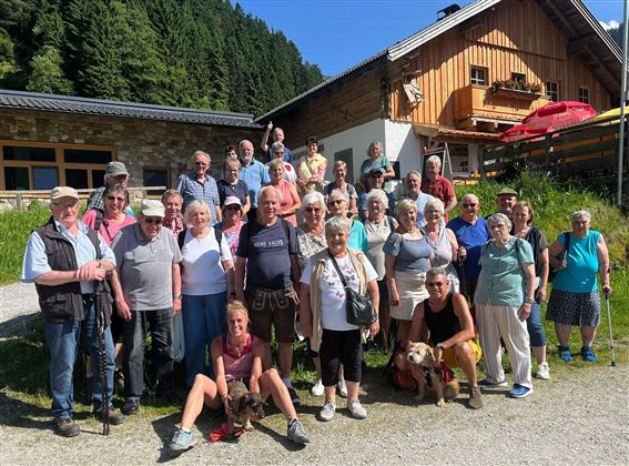 Eine große Gruppe von Menschen steht vor einem Holzhaus in den Bergen. Es ist ein sonniger Tag und einige Personen haben Hunde bei sich.