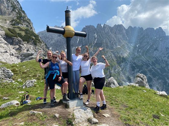A group of people stands in front of a mountain cross in the mountains. They are smiling and happily posing for the photo.