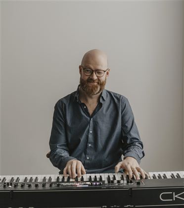 Ein lächelnder Mann sitzt am Klavier und spielt. Er trägt eine Brille und ein Hemd in blau.