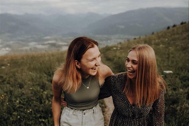 Zwei Frauen lachen und haben Spaß auf einem grünen Feld mit Bergen im Hintergrund. Die fröhliche Atmosphäre und die Natur schaffen eine positive Stimmung.