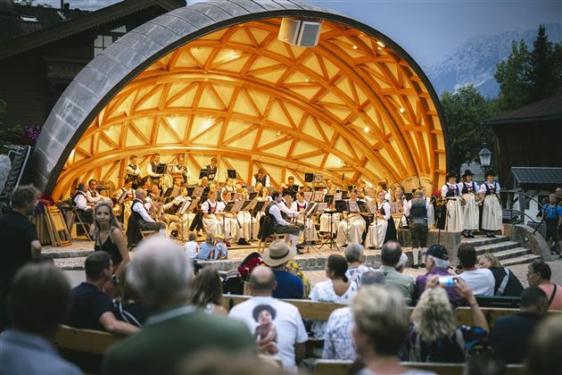 Ein Orchester spielt in einem einzigartigen Pavillon vor einem Publikum. Menschen sitzen auf Bänken und genießen das Konzert.